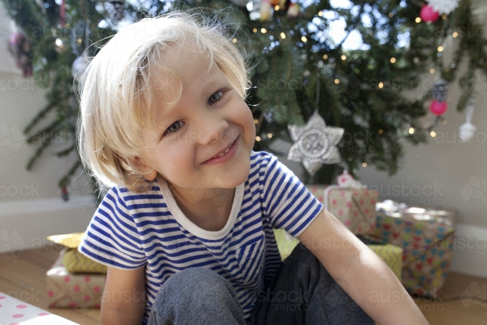 Young boy sitting in front of Christmas tree smiling - Australian Stock Image