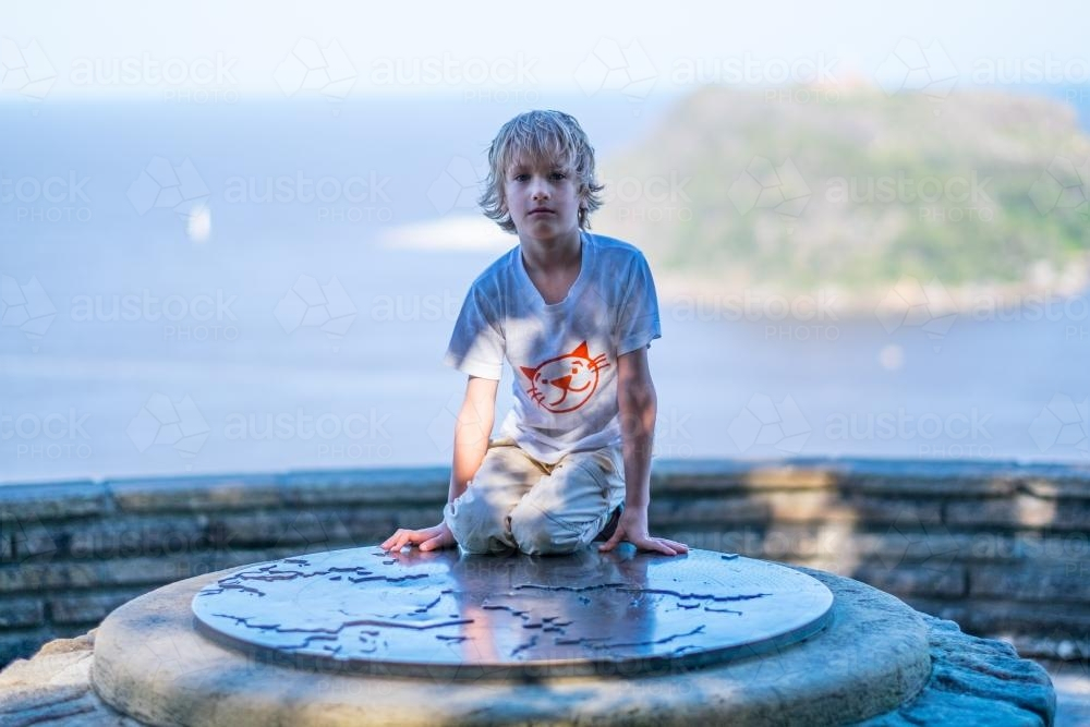 Young boy sitting on a circular stone structure with a map engraved on surface - Australian Stock Image