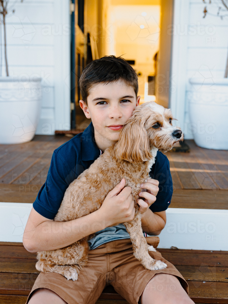 Young boy sitting on a wooden floor holding a light brown dog. - Australian Stock Image