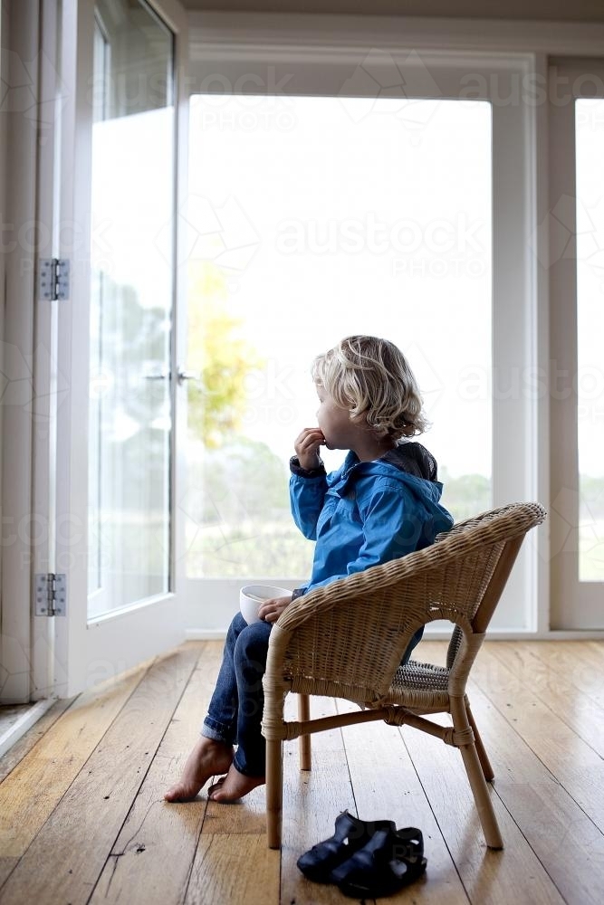 Young boy sitting on chair, eating and looking outside - Australian Stock Image