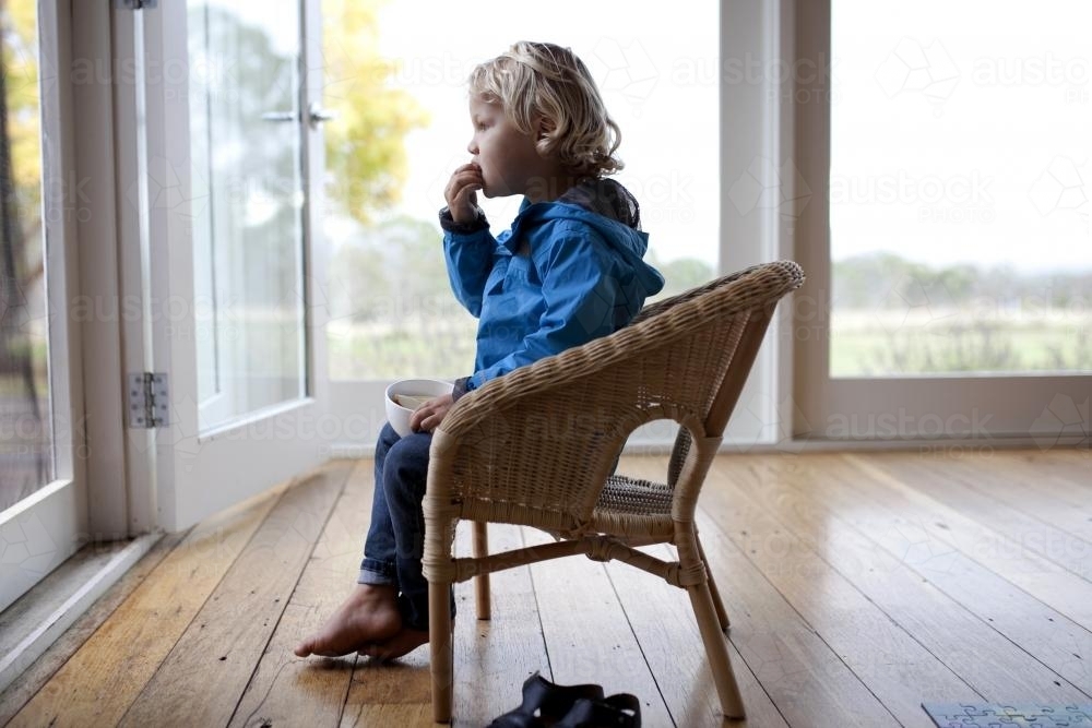 Young boy sitting on chair looking outside while eating - Australian Stock Image