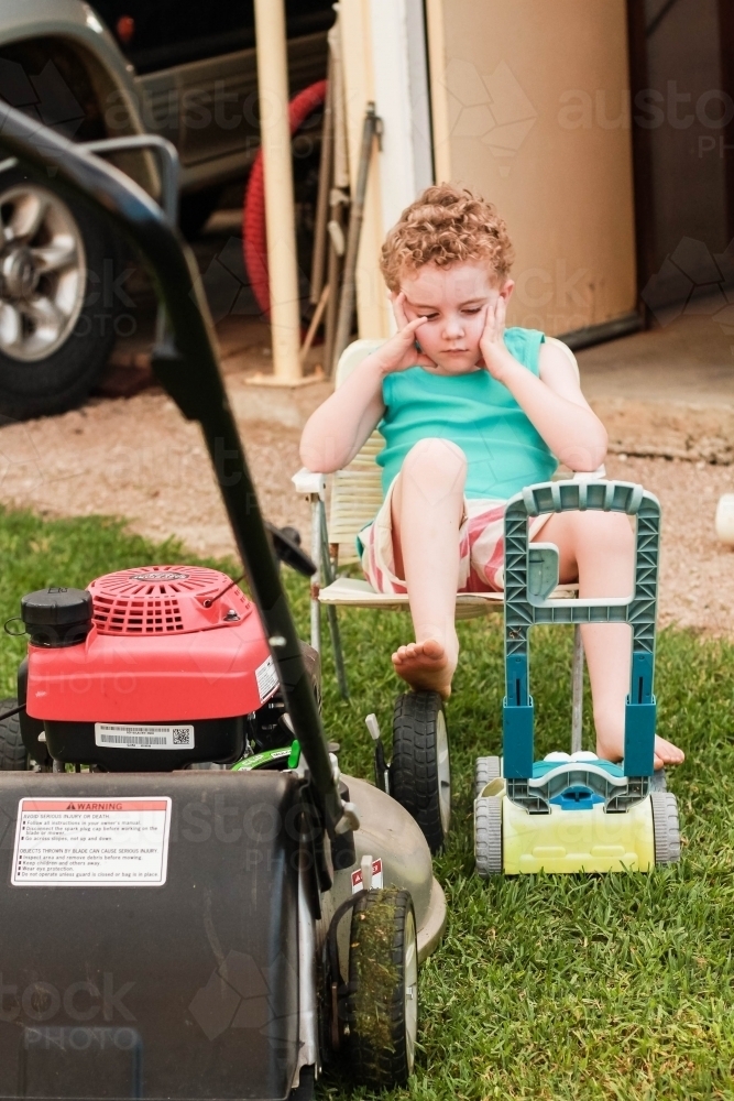 Young boy sitting on chair resting head with foot on lawn mower - Australian Stock Image
