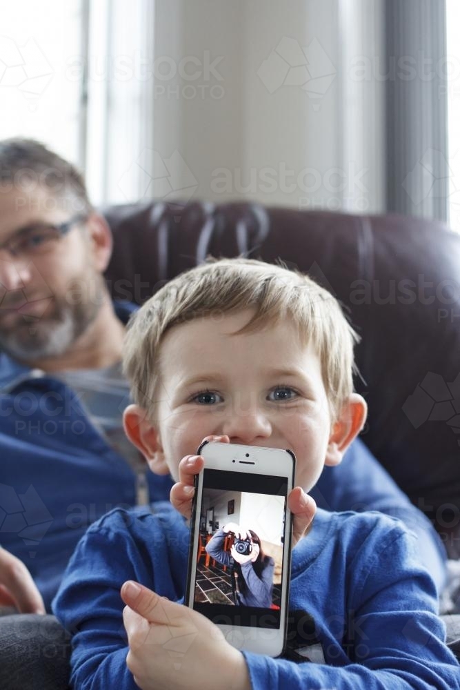 Young boy sitting on fathers lap holding phone with photo on screen - Australian Stock Image