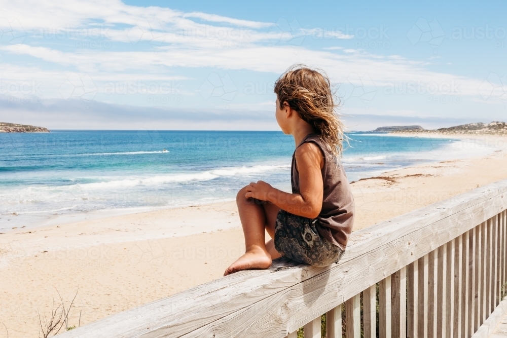 Young boy sitting on wooden fence looking at the ocean - Australian Stock Image