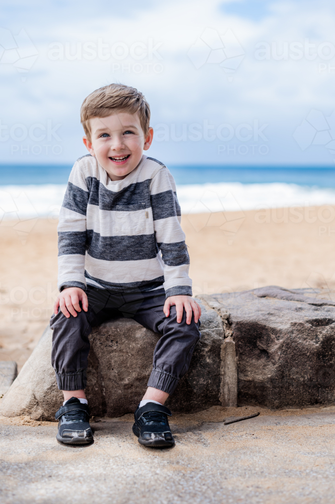 Young boy smiles while sitting on a rock near the beach with ocean in the background - Australian Stock Image