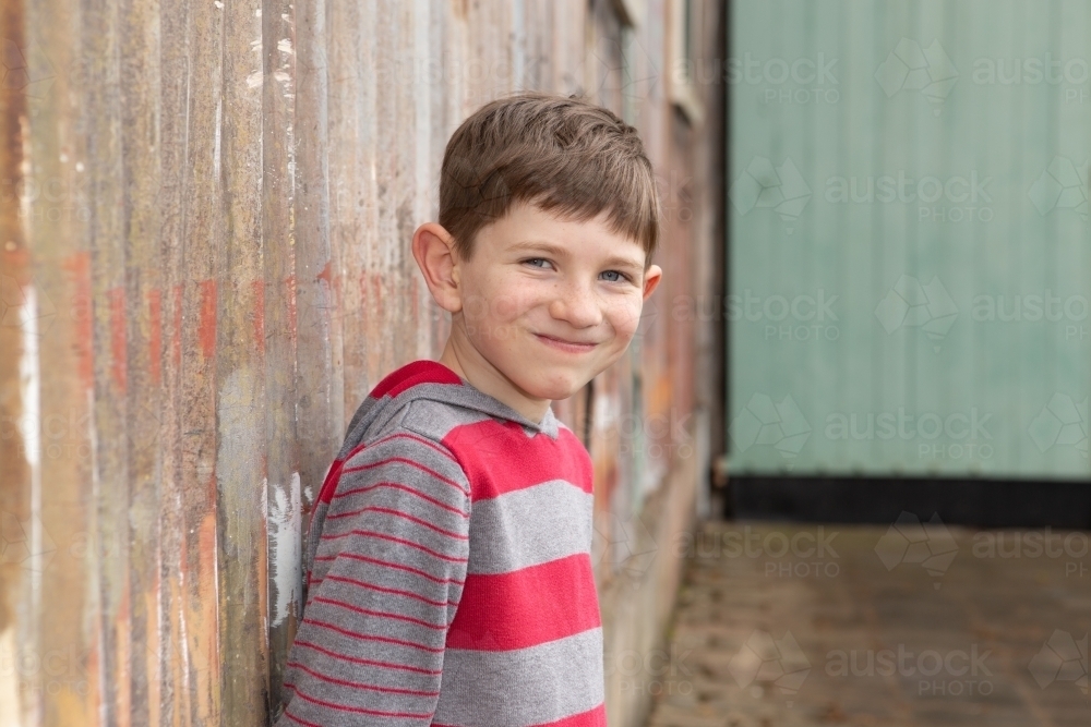 Young boy smiling at camera in front of rusty wall - Australian Stock Image