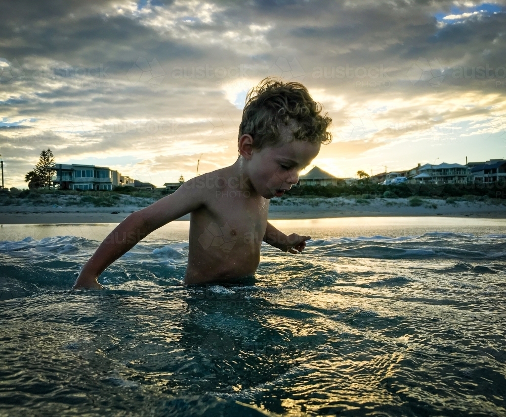 Young boy standing in ocean, looking down at sunrise. - Australian Stock Image