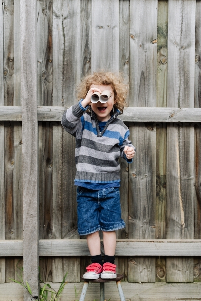 Young boy standing on a stool playing with cardboard toilet roll binoculars in the backyard - Australian Stock Image