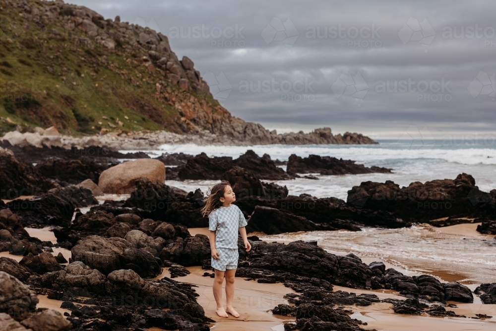 Young boy standing on beach between rocks with cliffs behind - Australian Stock Image