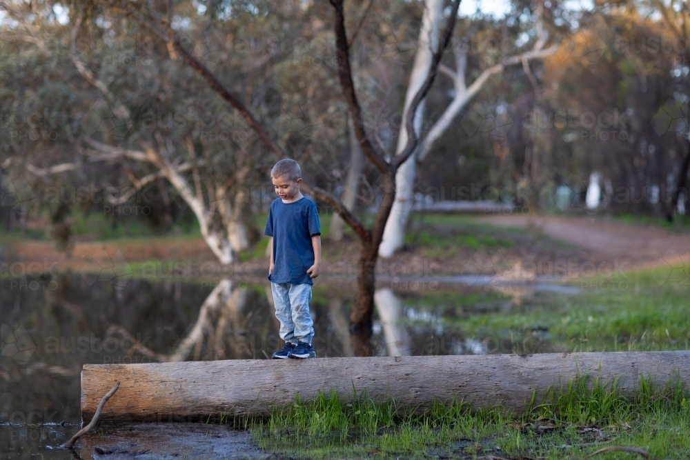 young boy standing on log on edge of lake - Australian Stock Image