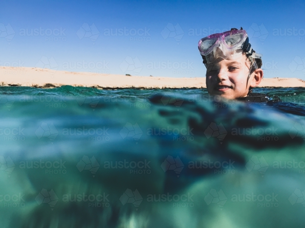 Young boy swimming in ocean with snorkelling mask on head looking at camera smiling - Australian Stock Image