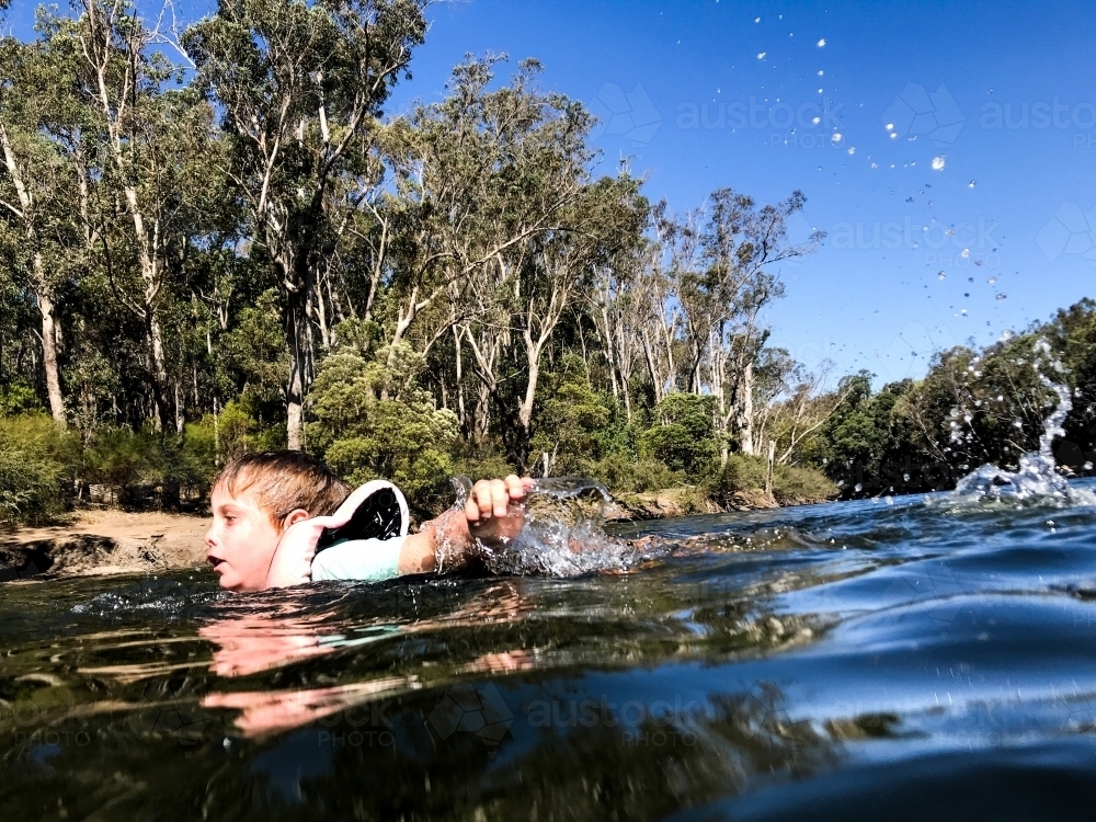 Young boy swimming in river with life jacket on and natural Forrest in background - Australian Stock Image