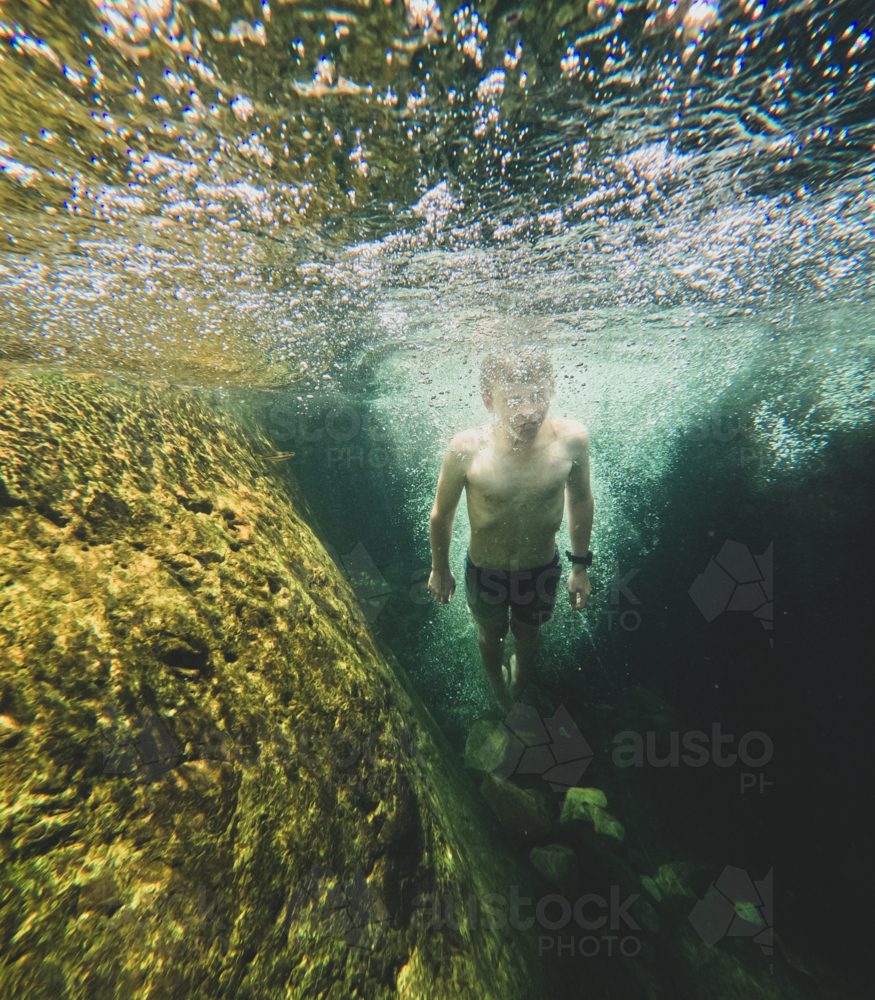 Young boy underwater surrounded by bubbles in clear dark green waters, rocks in background - Australian Stock Image