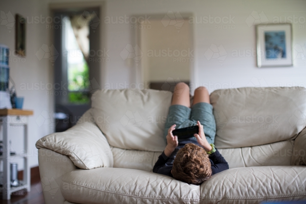 Young boy upside down on couch, playing a video games - Australian Stock Image