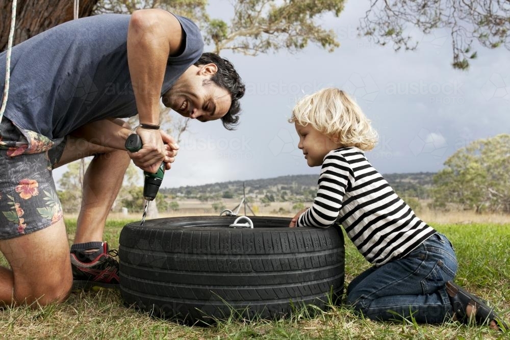 Young boy watching dad using power drill to make tyre swing - Australian Stock Image