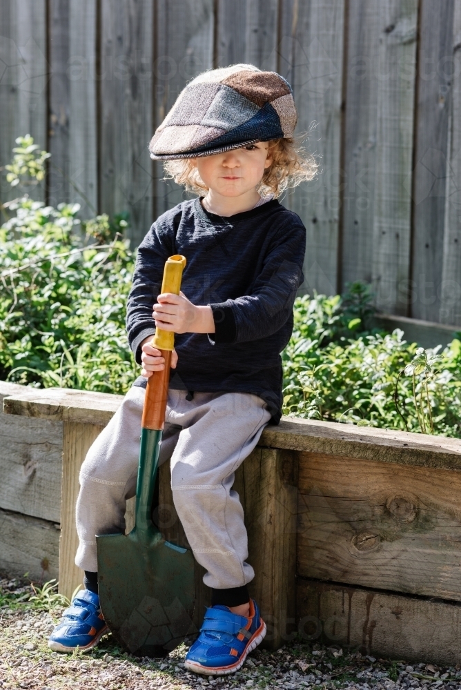 Young boy wearing a beret sitting with a shovel in the garden - Australian Stock Image