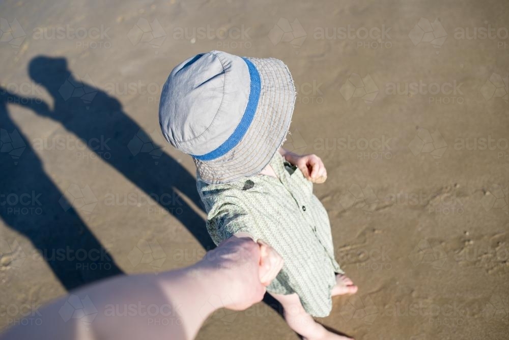 Young boy wearing a hat at the beach and holding hands seen from adult's POV - Australian Stock Image