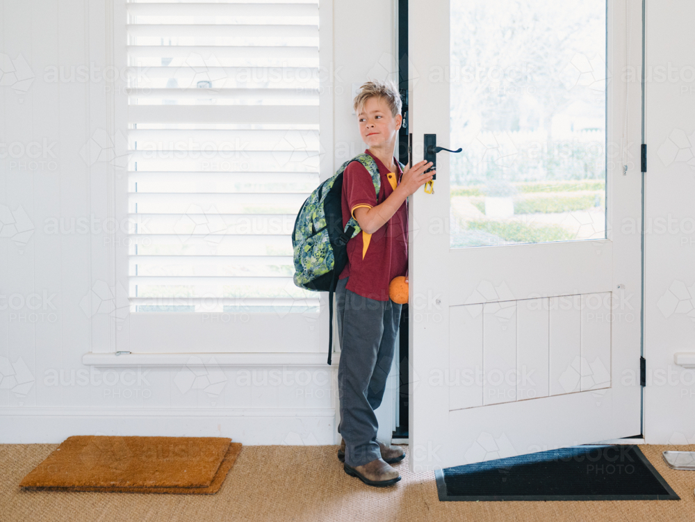 Young boy wearing backpack standing at the doorway holding a small orange ball - Australian Stock Image