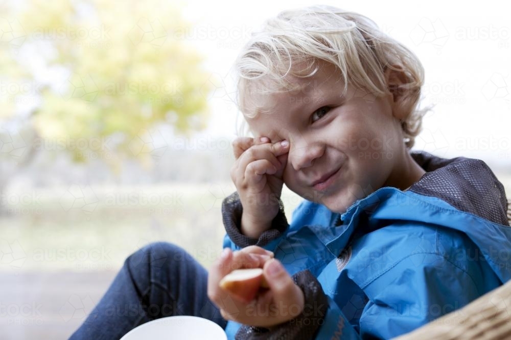Young boy wearing blue jacket eating apple looking at camera - Australian Stock Image