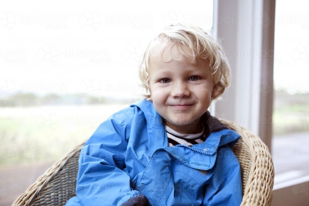 Young boy wearing blue jacket sitting in wicker chair in front of a window - Australian Stock Image