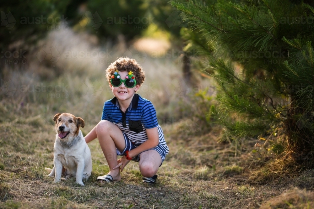 Young boy wearing glasses squatting next to dog in pine trees - Australian Stock Image
