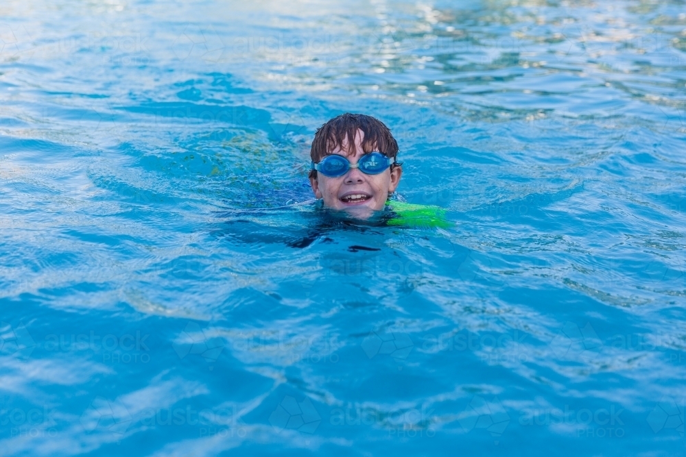 Young boy wearing goggles smiling happy swimming in pool - Australian Stock Image