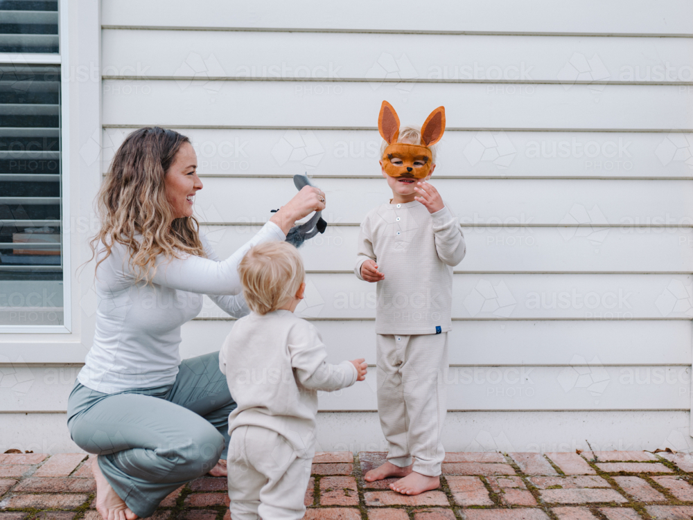 Young boy wearing kangaroo mask while mum crouching and holding koala mask for toddler brother - Australian Stock Image