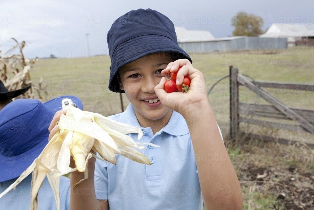 Young boy wearing school uniform holding up freshly picked vegetables - Australian Stock Image