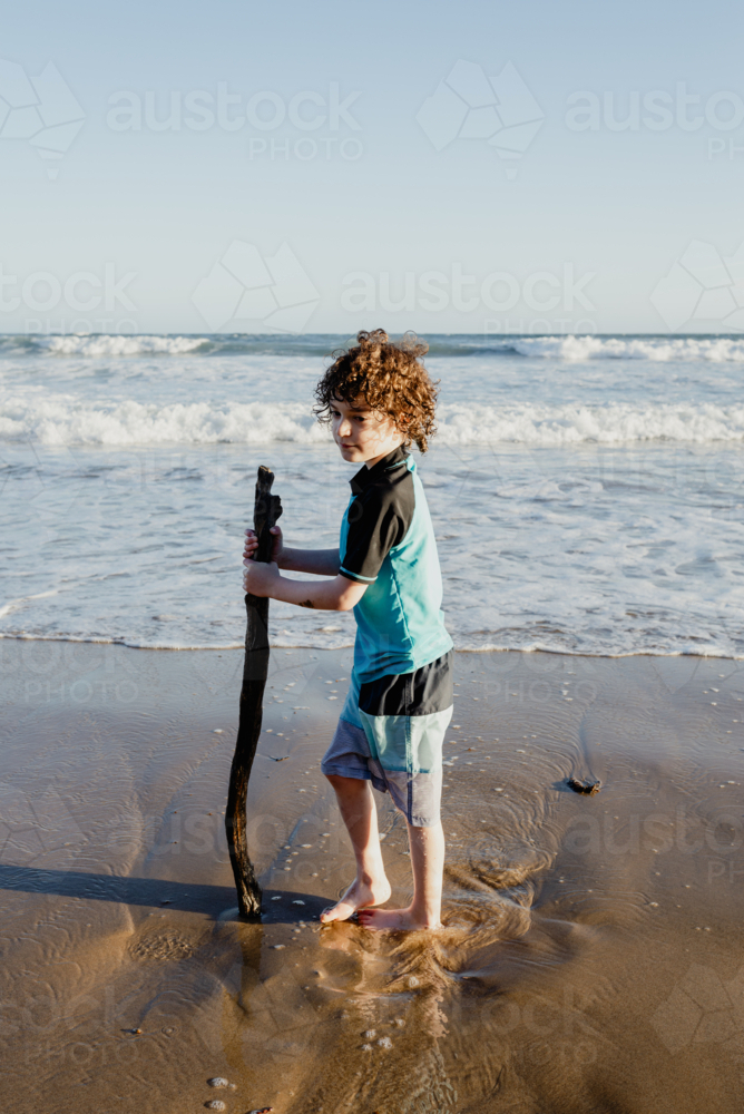 Young boy with curly hair holding a large stick standing on a beach in the afternoon - Australian Stock Image