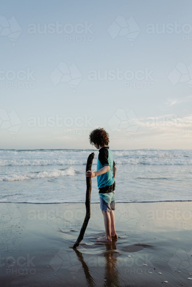 Young boy with curly hair holding a large stick standing on a beach in the afternoon - Australian Stock Image