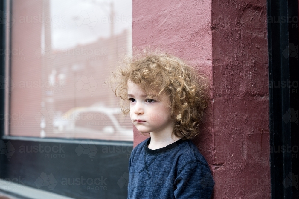Young boy with curly hair standing against a pink brick wall - Australian Stock Image