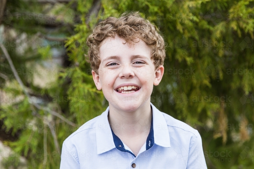Young boy with curly hair standing in front of tree smiling - Australian Stock Image