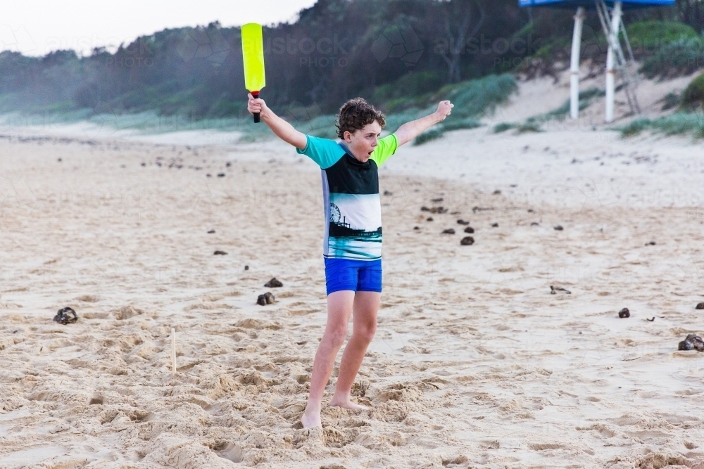 Young boy with fists raised in the air holding cricket bat excited on beach - Australian Stock Image