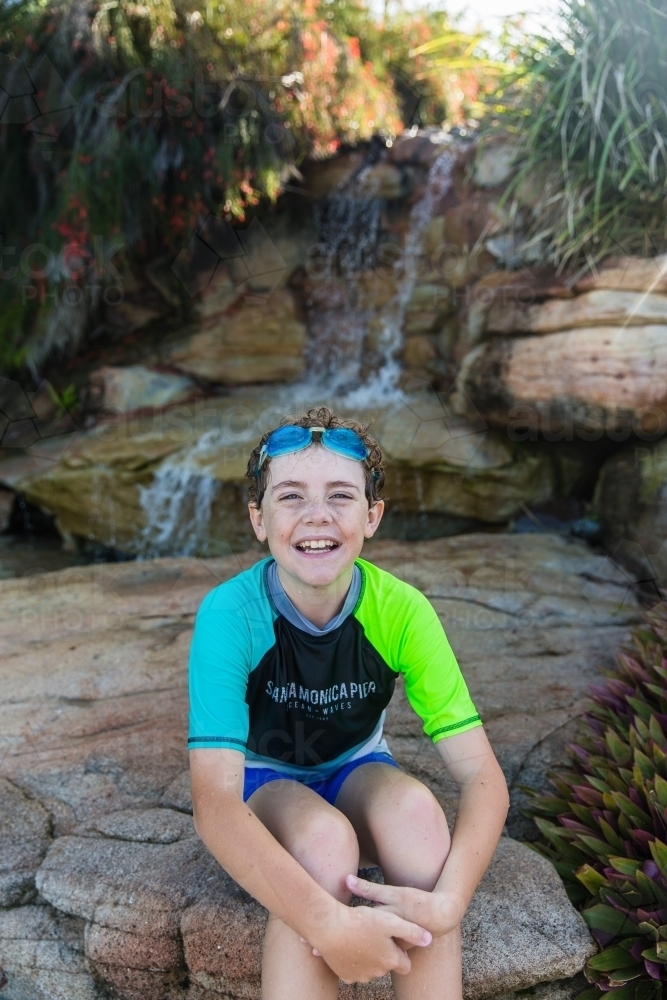 Young boy with goggles sitting on rocks in front of waterfall happy smiling - Australian Stock Image