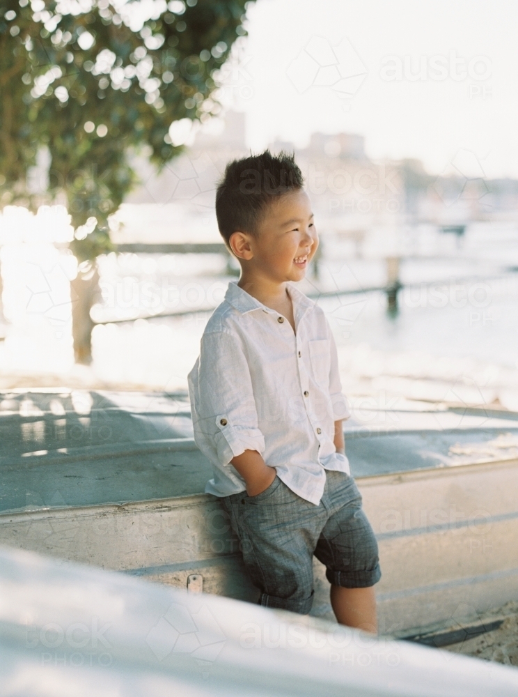 Young boy with his hands in his pockets, leaning against an upturned boat by the water - Australian Stock Image