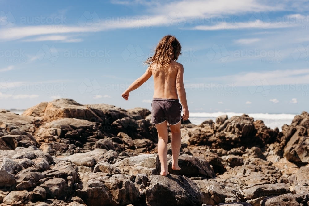 Young boy with long hair walking on rocks in ocean - Australian Stock Image