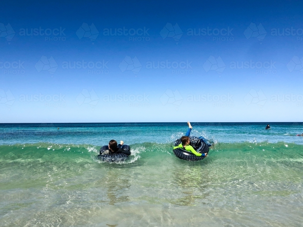 young boys in wetsuits lying on inflatable tyres on small beach waves - Australian Stock Image