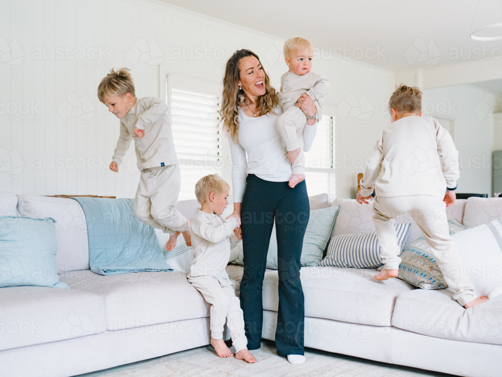 Young boys playing and jumping on the couch with mum carrying the baby - Australian Stock Image