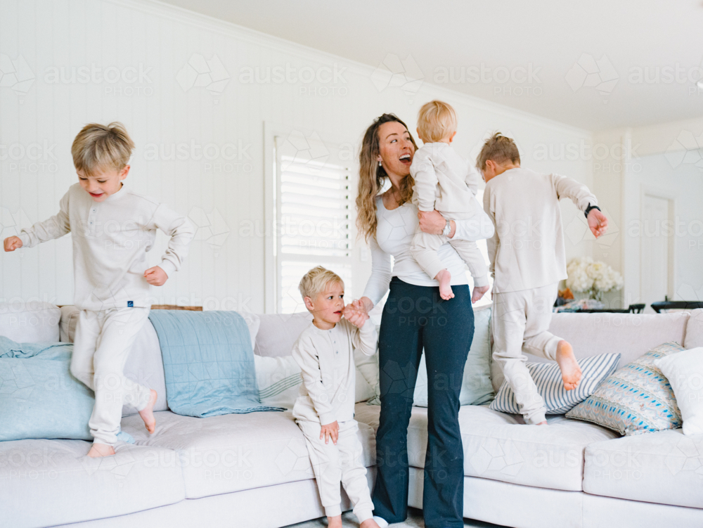 Young boys playing and jumping on the couch with mum carrying the baby - Australian Stock Image
