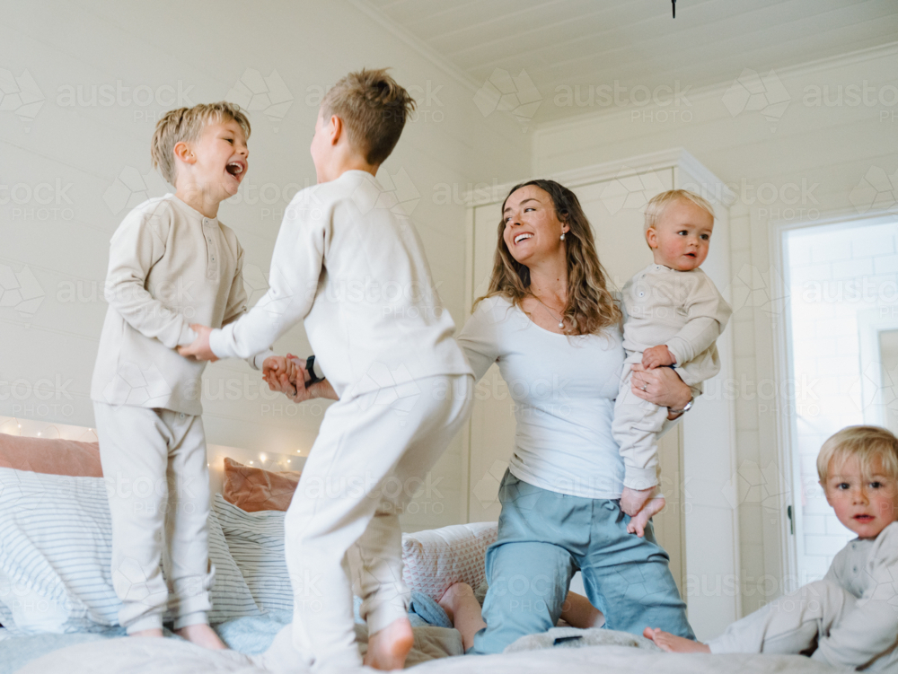 Young boys playing on the bed jumping with mum holding baby brother - Australian Stock Image