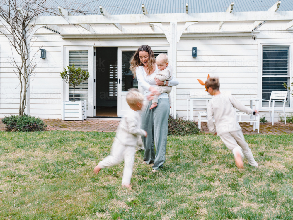 Young boys running around mum holding a baby in the garden - Australian Stock Image