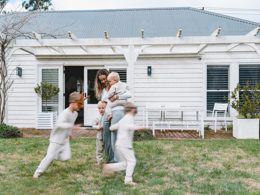 Young boys running around mum holding a baby in the garden - Australian Stock Image