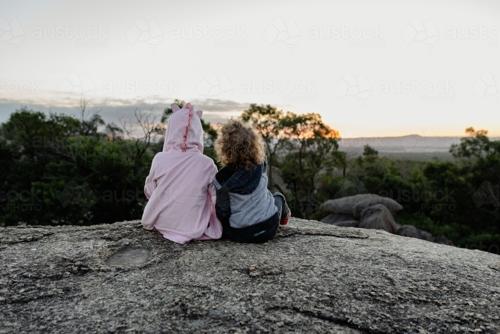 Young brother and sister sitting together on a rocky ledge in the Australian bush watching a sunset - Australian Stock Image