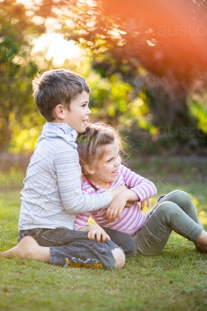 Young brother and sister wrestling on the lawn outside in afternoon light - Australian Stock Image
