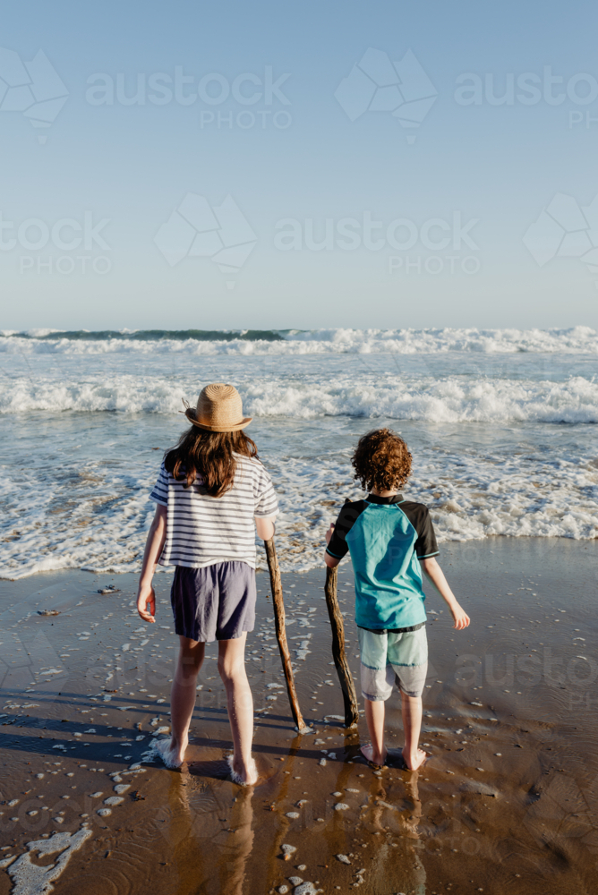 Young brother & sister (young boy and girl) holding sticks standing together at the beach - Australian Stock Image