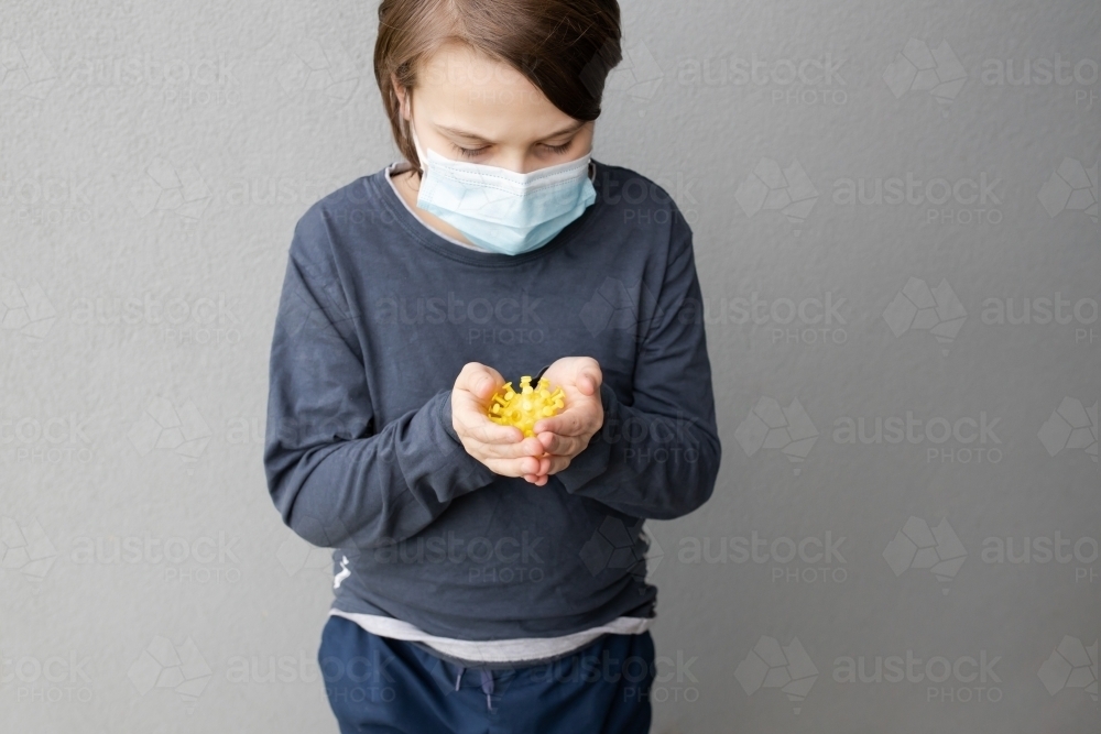 Young caucasian boy wearing a blue surgical mask and holding a corona virus model during the COVID-1 - Australian Stock Image