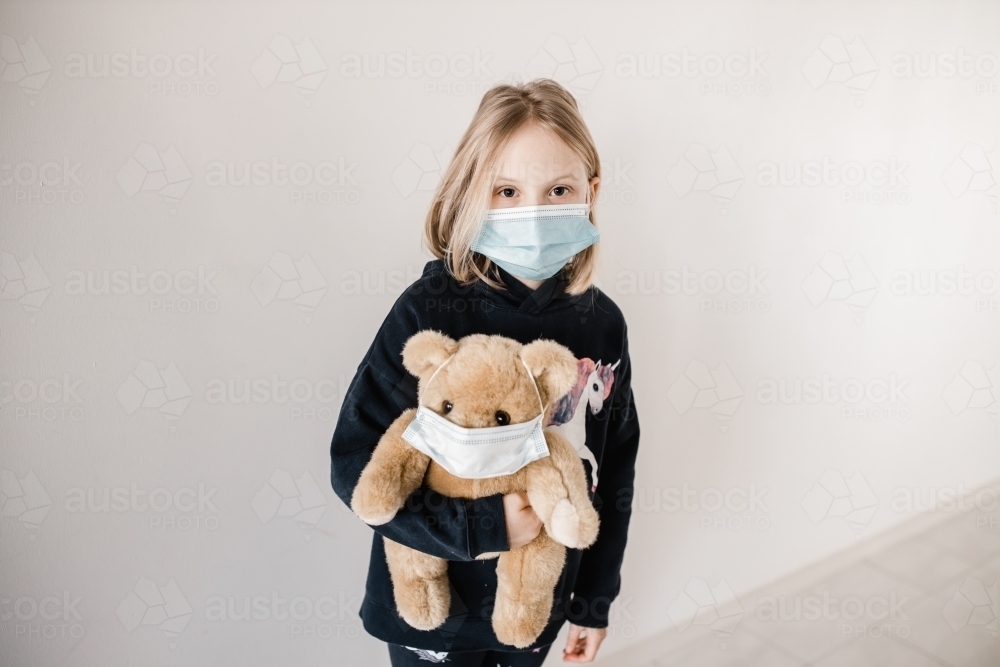 Young caucasion girl wearing a mask and holding a teddy bear who is also wearing a surgical mask dur - Australian Stock Image