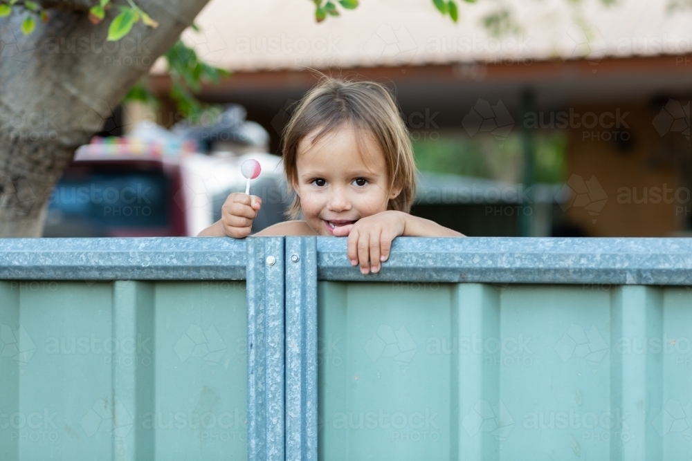 young child holding lollipop and peeking over a fence - Australian Stock Image