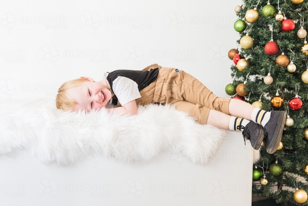 Young child lying on white rug laughing with Christmas tree - Australian Stock Image
