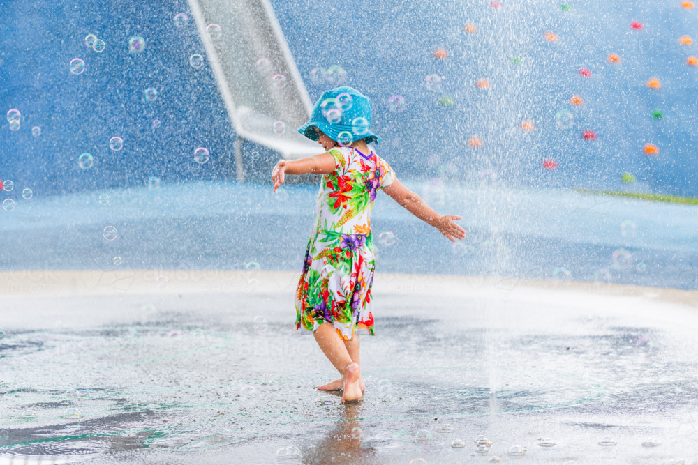 Young child running barefoot through water fountain play area at park in swim hat and dress - Australian Stock Image